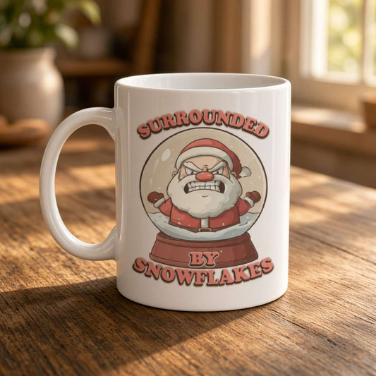 White ceramic mug on a sunlit wooden table, printed with an angry cartoon Santa in a snow globe and the words “Surrounded by snowflakes”; playful holiday humor.