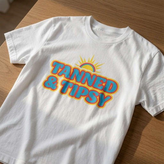 White Tanned & Tipsy t-shirt laid flat on a wooden surface showing the full retro sun design, fun ladies summer slogan shirt