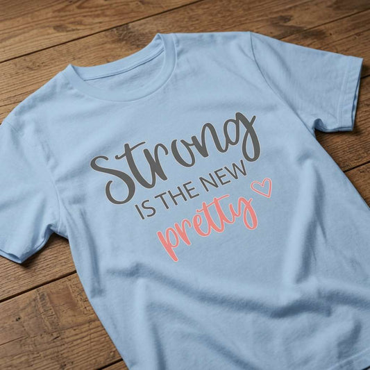 Blue Strong is the New Pretty t-shirt laid flat on a wooden surface showing the full script lettering and heart, fun ladies motivational strong statement tee that every no-nonsense lady will claim