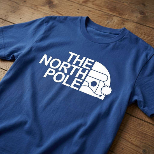 Blue The North Pole t-shirt laid flat on a wooden surface showing the full logo design, fun and witty xmas day shirt that every cheeky lad will claim
