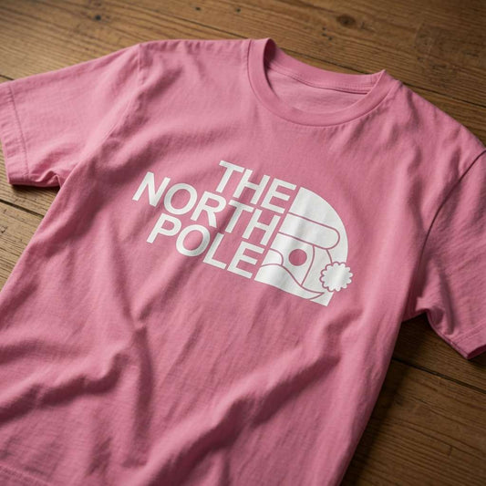 Pink The North Pole t-shirt laid flat on a wooden surface showing the full logo design, fun and humorous Santa xmas pub shirt