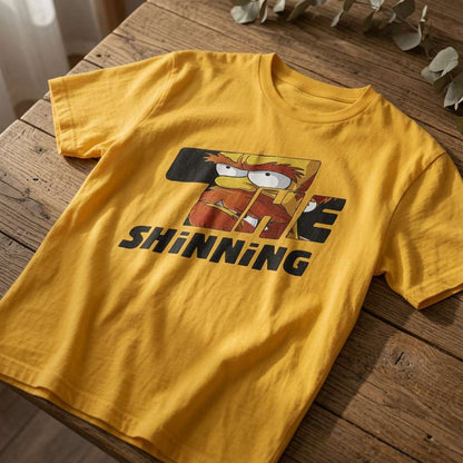 Yellow The Shinning t-shirt laid flat on a wooden surface showing the full cartoon graphic, mens humorous inspired by horror tee