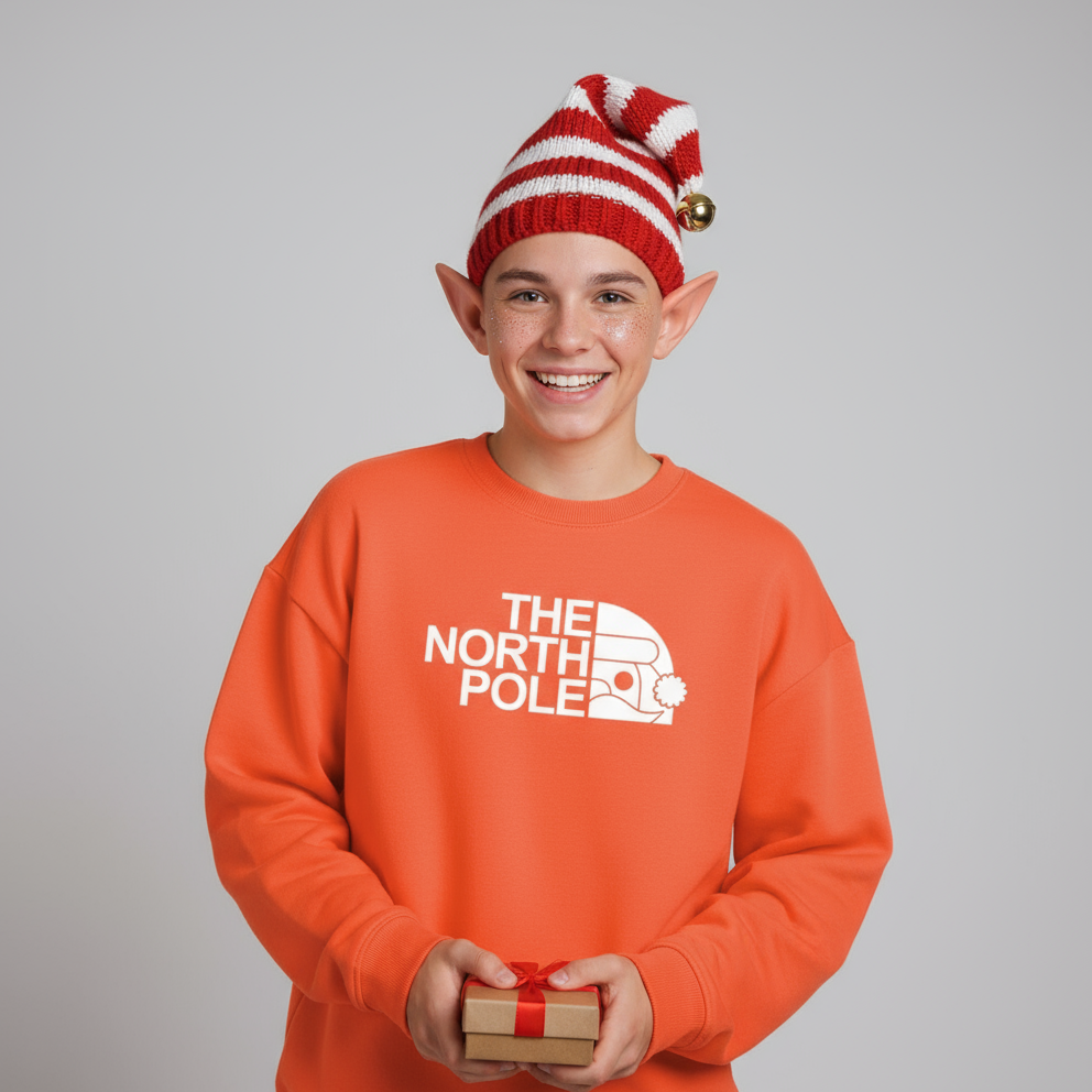 An elf wearing an orange 'The North Pole' Christmas sweatshirt and a red and white striped elf hat, holding a small gift box. 
