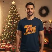 Man wearing a navy blue Christmas Turkey t-shirt with 'The Gobbling' design in a festive room with Christmas tree and fireplace.