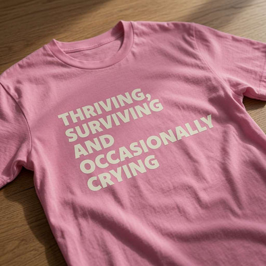 Pink Thriving, Surviving and Occasionally Crying t-shirt laid flat on a wooden surface showing the full slogan, cheeky witty and relatable ladies design that every no-nonsense lady will claim