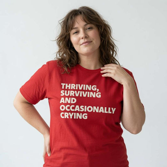 Woman with one hand on her shoulder while wearing a red Thriving, Surviving and Occasionally Crying t-shirt, funny cheeky and witty relatable ladies text shirt
