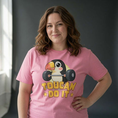 Woman smiling with one hand on her hip while wearing a pink Toucan Do It t-shirt featuring a cartoon toucan lifting weights, cheeky pun and motivational bird ladies shirt