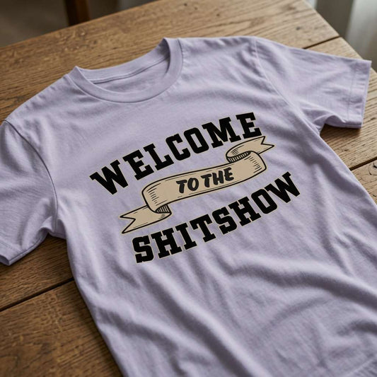 Lavender Welcome to the Shitshow t-shirt laid flat on a wooden surface showing the full banner slogan, fun relatable famous slogan ladies shirt full of pure attitude