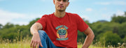 A Man sits on grass in a park, wearing a red funny t-shirt with a cartoon succulent and text 
