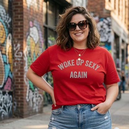 Woman smiling with one hand in her pocket and sunglasses on while wearing a red Woke Up Sexy Again t-shirt outdoors, funny witty ladies quote design