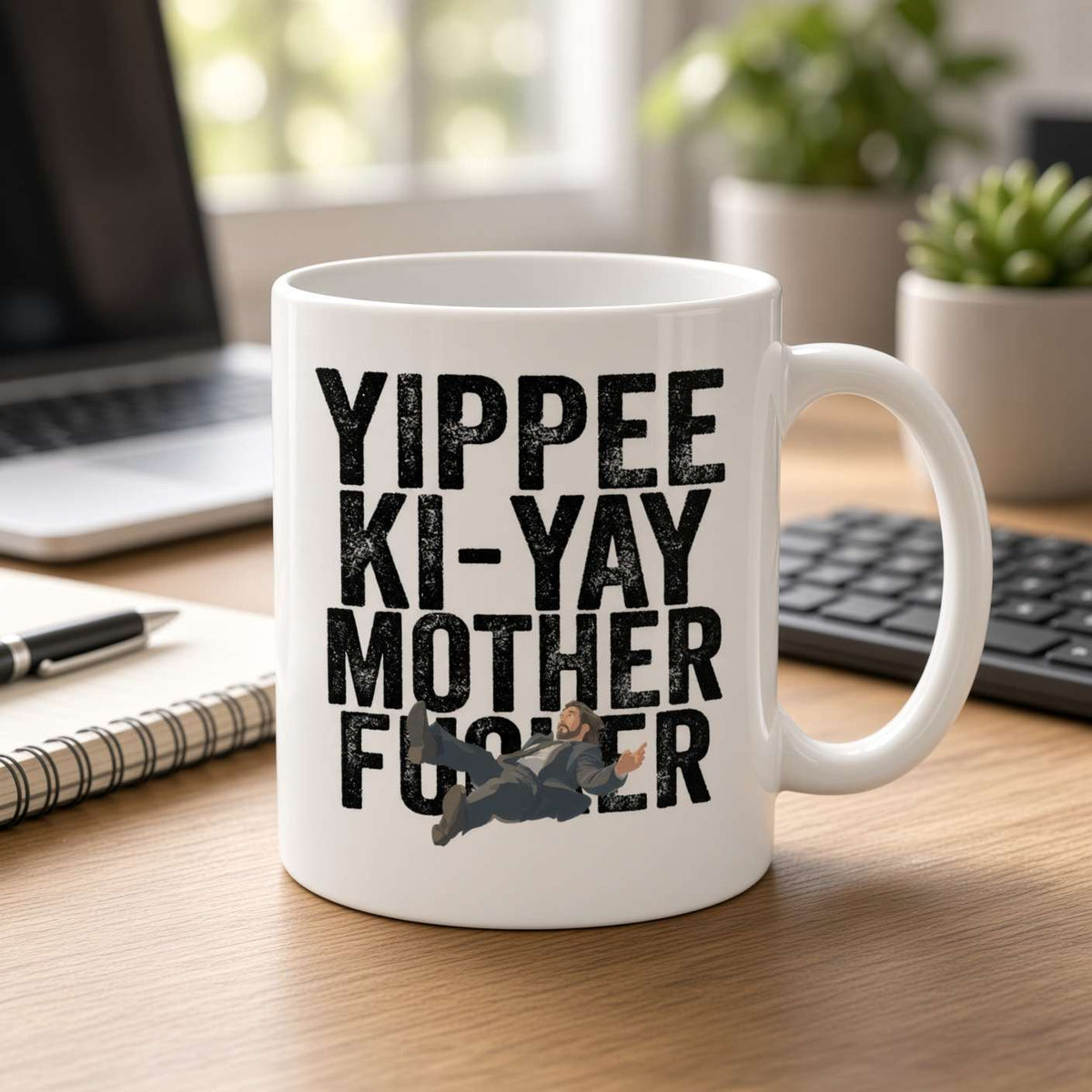 White coffee mug with bold “Yippee ki-yay, motherf***er” lettering and a small action-hero illustration sits on a bright, tidy office desk.