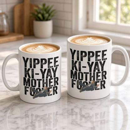 Two white latte mugs with leaf-art foam and bold “Yippee Ki-Yay Mother F…” lettering sit on a marble counter, creating a playful kitchen scene.
