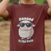 Maroon funny t-shirt with a cool cartoon sheep wearing sunglasses and slogan 'Baaaad To The Bone' on a blurred natural background.