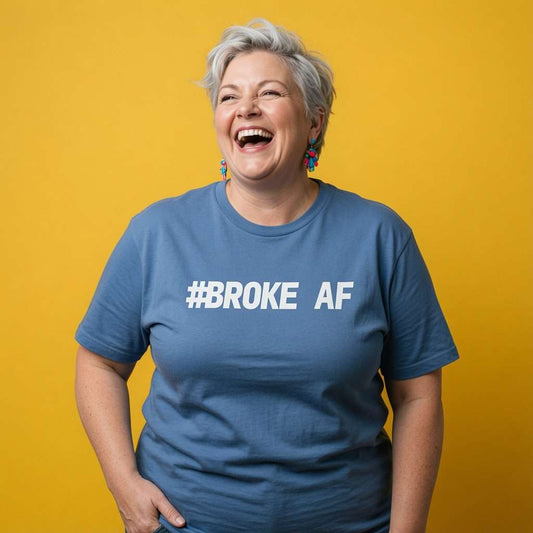 A laughing, gray-haired woman in colorful earrings wears a blue “#BROKE AF” T-shirt against a bright yellow background, conveying joyful humor.