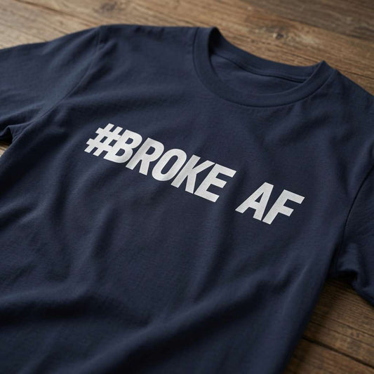 A navy T-shirt lies on a wooden surface, printed with bold white “#BROKE AF” lettering; the casual design conveys a blunt, humorous tone.
