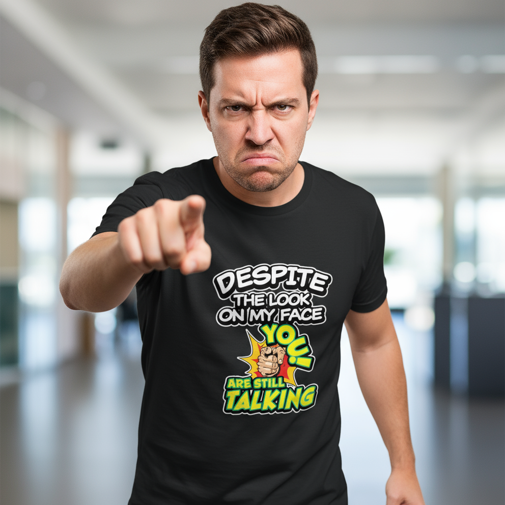 Man with an angry expression points directly at the camera. He wears a black shirt with bold text that reads, "Despite the look on my face, you are still talking." The background is a blurred indoor setting.