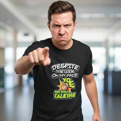 Man with an angry expression points directly at the camera. He wears a black shirt with bold text that reads, "Despite the look on my face, you are still talking." The background is a blurred indoor setting.