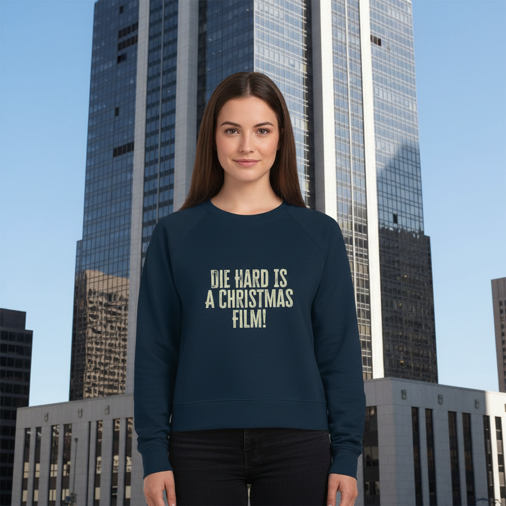 A woman stands smiling in front of a tall modern skyscraper, wearing a navy sweatshirt with "Die Hard is a Christmas film" printed on it. The tone is playful.