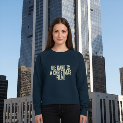 A woman stands smiling in front of a tall modern skyscraper, wearing a navy sweatshirt with "Die Hard is a Christmas film" printed on it. The tone is playful.