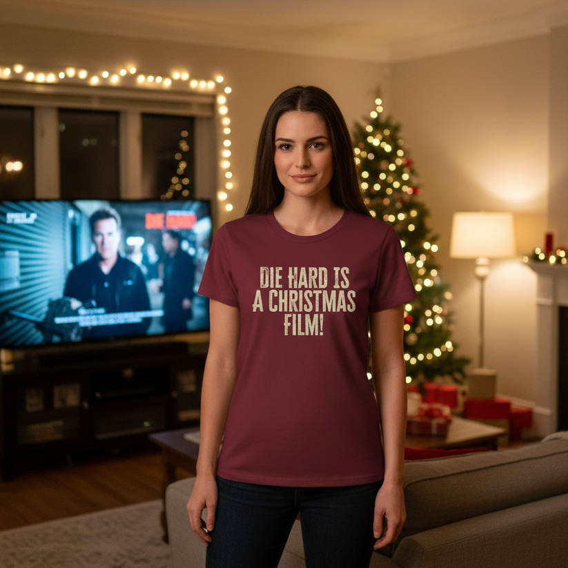 A woman stands in a cozy, festive living room wearing a maroon shirt with "Die Hard is a Christmas Film!" text. A Christmas tree and lights add warmth.