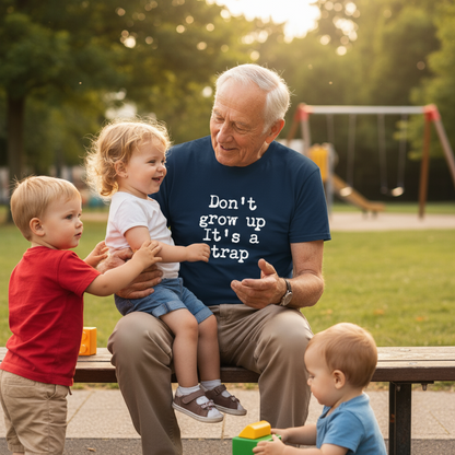Man sitting on a bench with three children in a park, wearing a shirt with the text 'don't grow up it's a trap'