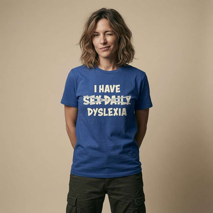 A woman with shoulder-length brown hair stands against a plain beige studio background wearing a blue short-sleeve T-shirt with white text that reads 'I HAVE SEX DAILY DYSLEXIA' and dark cargo pants.