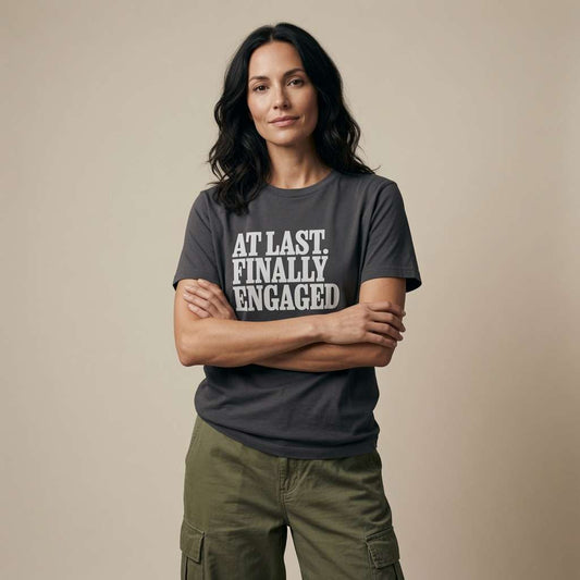 A woman stands with arms crossed wearing a dark ink gray graphic t-shirt that reads 'AT LAST FINALLY ENGAGED' in bold white text, paired with olive green cargo pants, against a solid beige studio background.
