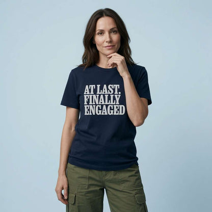 A woman wearing a navy blue crewneck t-shirt with the text 'AT LAST, FINALLY ENGAGED' printed in bold white capital letters, paired with olive green cargo pants, set against a solid light blue background.