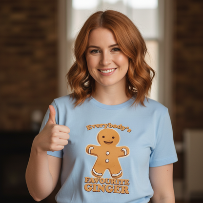Young woman with red hair smiling, wearing a blue shirt with a gingerbread man and the text "Everybody's Favourite Ginger." She gives a thumbs up.