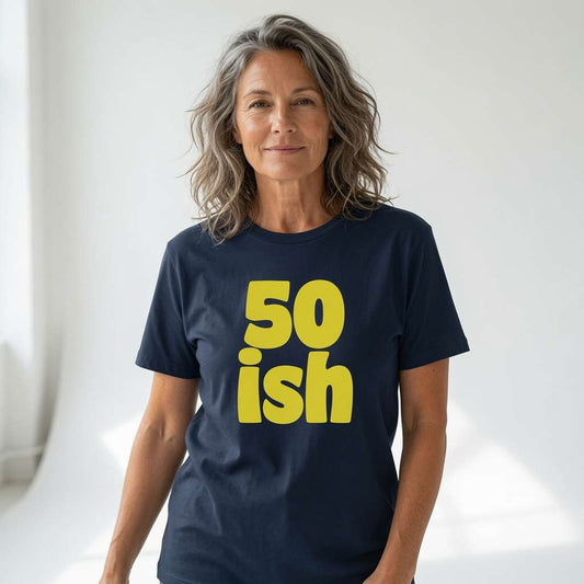 A woman with shoulder-length gray-blonde hair wearing a navy blue t-shirt with the words '50 ish' printed in bold yellow letters on the front, set against a bright, softly lit white studio background.