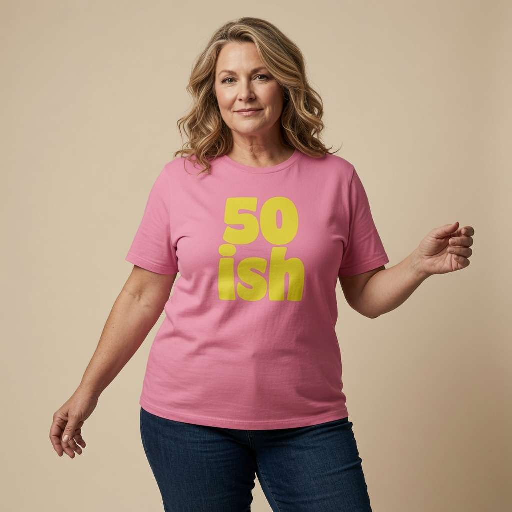 A woman modeling a pink short-sleeve t-shirt with '50 ish' printed in bold yellow text on the front, paired with dark blue jeans against a solid beige background.