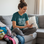 Woman sitting on a couch with clothes and using a phone, wearing a green t-shirt with text.