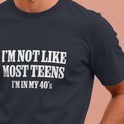 Black funny dad slogan t-shirt with white text 'I'M NOT LIKE MOST TEENS I'M IN MY 40'S' on a beige background