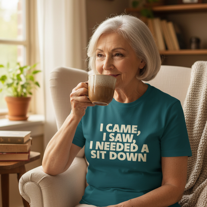 Woman sitting in a cozy room, drinking coffee and wearing a teal t-shirt with humorous text.