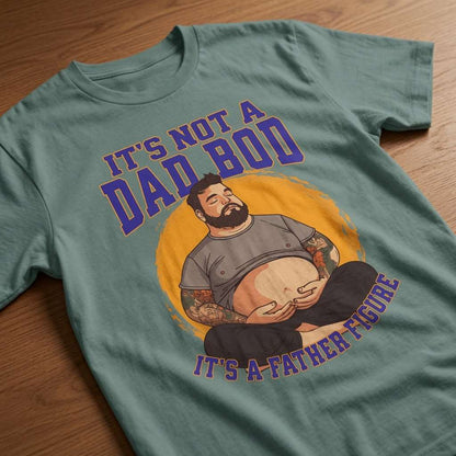 Sage green t-shirt laid flat on a wooden surface showing the full cartoon dad illustration and the slogan “It’s Not a Dad Bod, It’s a Father Figure”