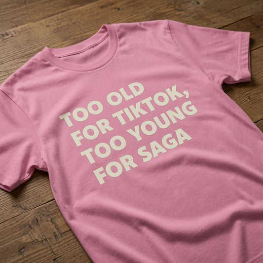 Pink t-shirt laid flat on a wooden surface with the white slogan “Too Old For TikTok, Too Young For Saga”, humorous age-related banter for ladies