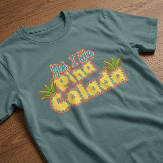 Green bay Yes, I Like Pina Colada t-shirt laid flat on a wooden surface showing the colourful cocktail slogan with leaves, pina colada cocktail slogan design full of pure attitude