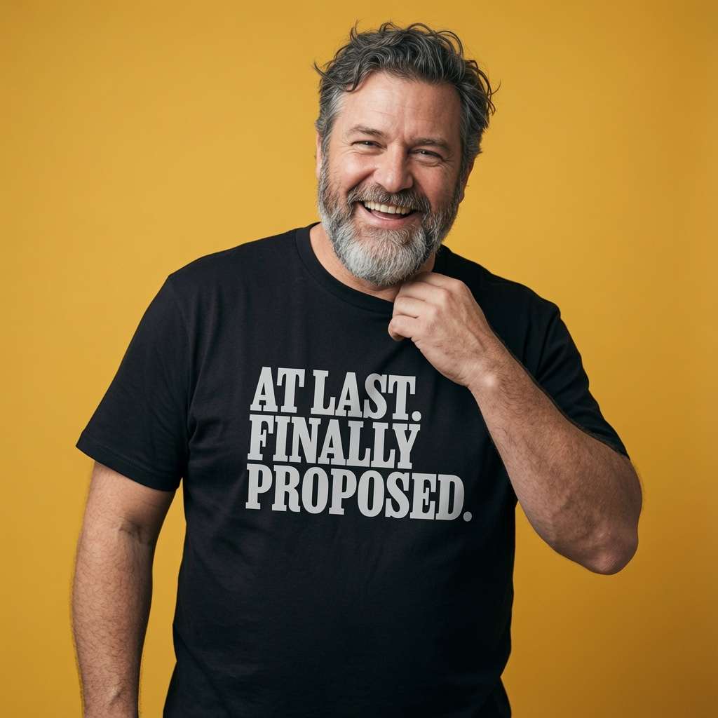 Bearded man smiling with one hand near his chin while wearing a black At Last. Finally Proposed. t-shirt, mens couples matching engagement text full of pure relief