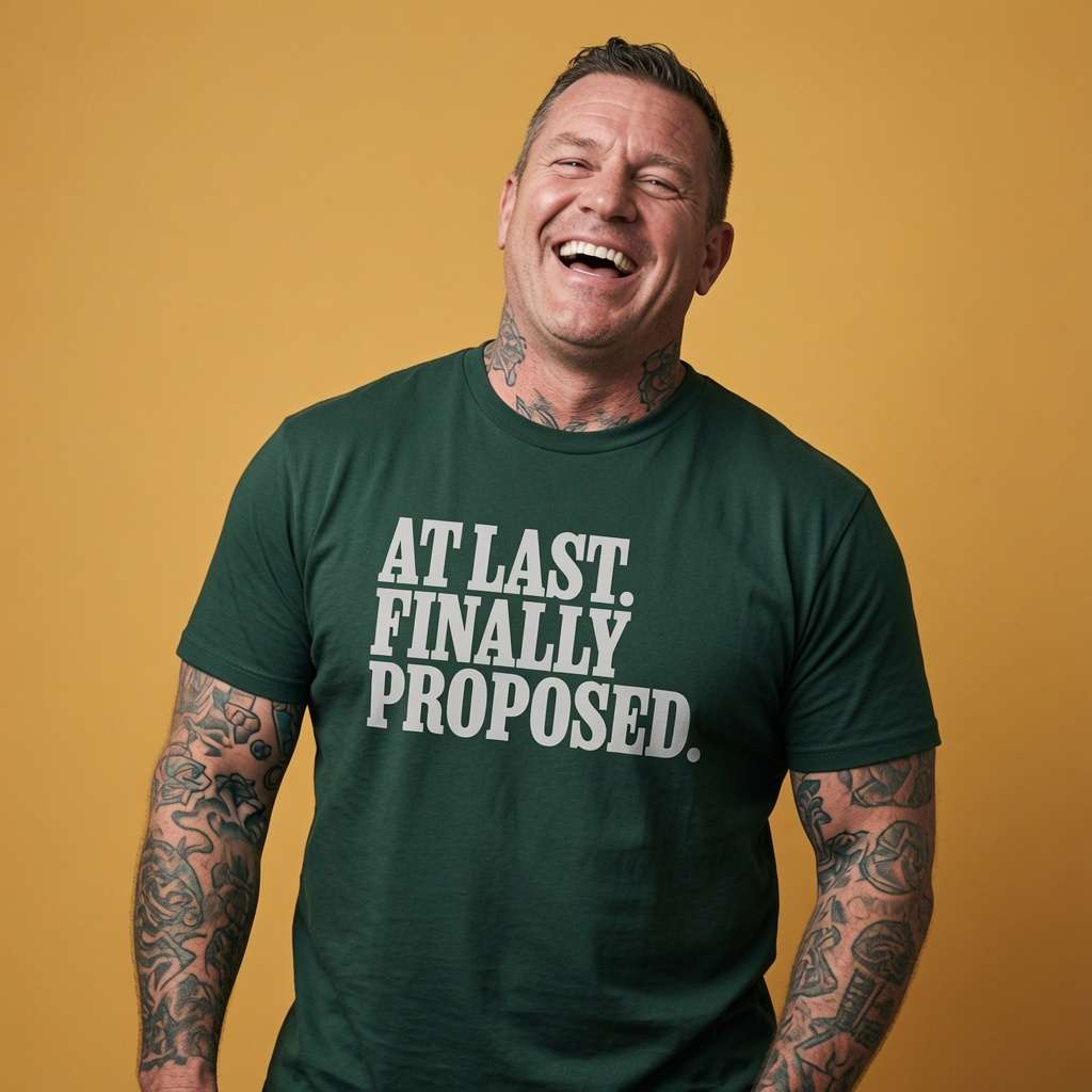 Tattooed man laughing in a dark green At Last. Finally Proposed. t-shirt, witty matching couples engagement slogan that every newly engaged man will claim