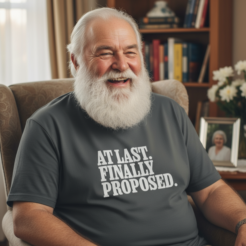 Man wearing a t-shirt with 'AT LAST. FINALLY PROPOSED.' text, sitting in a room with books and a photo frame.