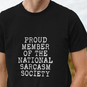 Black sarcastic slogan t-shirt with white text 'PROUD MEMBER OF THE NATIONAL SARCASM SOCIETY' on a blurred background