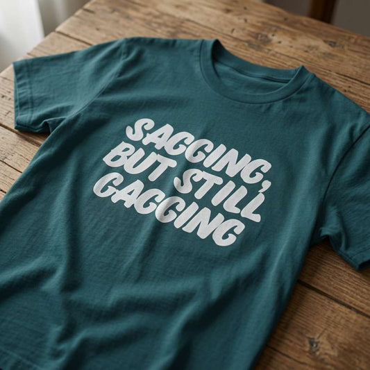 Teal green Sagging, But Still Gagging t-shirt laid flat on a wooden surface with bold white text, fun and rude ladies cheeky offensive gift slogan