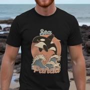 Person wearing a black t-shirt with a 'Sea Panda' design featuring an orca and waves, standing on a rocky beach.