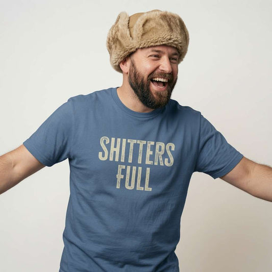Bearded man in a fur hat laughing with arms outstretched in a blue Shitters Full t-shirt, fun mens offensive xmas movie slogan that every Christmas Vacation fan will claim