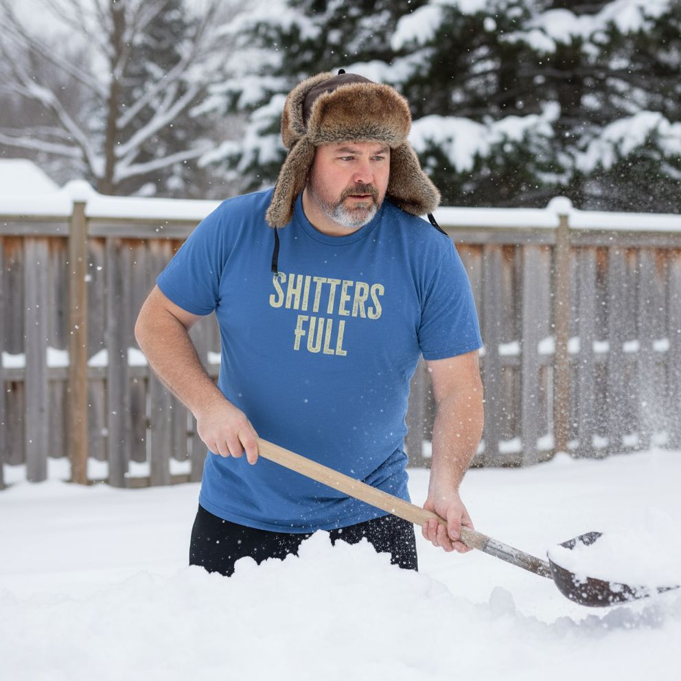 Man shoveling snow wearing a blue t-shirt with text and a fur hat in a snowy backyard.