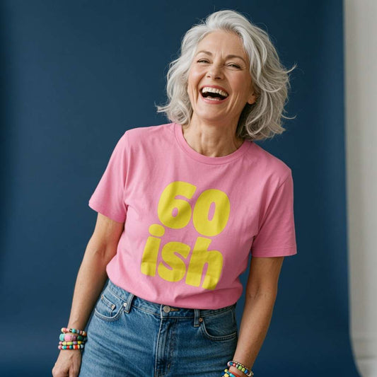 Grey-haired woman laughing while wearing a pink 60 ish t-shirt in bold yellow lettering, cheeky ladies 60th birthday slogan gift