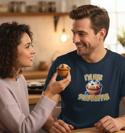 Man wearing a shirt with a cupcake design and text, holding a muffin in a kitchen setting.