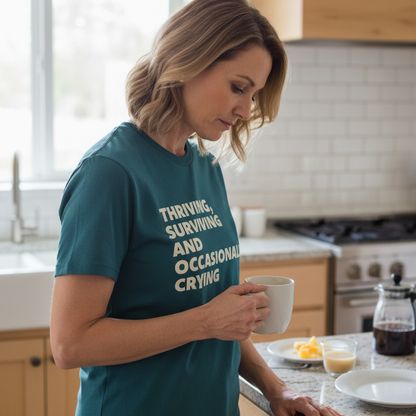 Woman in a kitchen wearing a teal t-shirt with text, holding a mug.