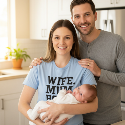 Woman holding a baby while standing next to a man in a kitchen, with the woman wearing a 'WIFE. MUM. BOSS' shirt.